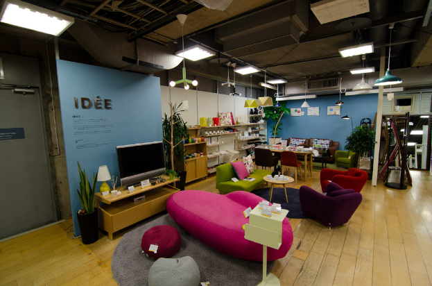 Eine Ladeneinrichtung mit Haushaltsgeräten und Möbeln, darunter ein Fernseher auf einem Tisch, eine pinkfarbene Couch, ein Sofa, zwei Stühle um einen kleinen Hocker, ein Esstisch mit Stühlen, ein großes Sofa im Hintergrund und verschiedene Gegenstände auf Regalen, alles beleuchtet von Deckenlampen.