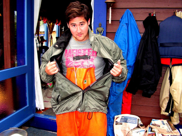 Eine Jacke und ein T-Shirt sind in der Mitte zu sehen, umgeben von verschiedenen Gegenständen wie Jacken, Zeitungen, einem Haus, Vorhängen, einer Tür, Fenstern, einer Glocke und einer Wand.