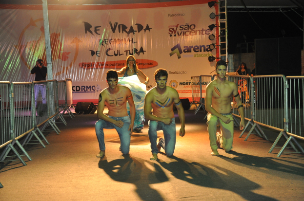 Drei shirtlose Jungs knien auf dem Boden während eines kulturellen Ereignisses, mit Grills auf beiden Seiten und einem großen Banner im Hintergrund, während eine Person auf der linken Seite unter Überkopfbeleuchtung ein Foto macht.