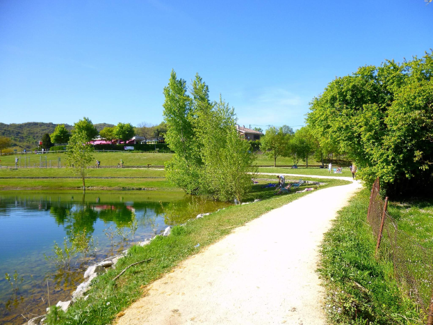 Ein Schotterweg neben einem Gewässer, gesäumt von grünem Gras, Pflanzen und Bäumen, mit einem Zaun daneben, Gebäuden, Hügeln und einem klaren blauen Himmel im Hintergrund, wo Menschen die Landschaft genießen.