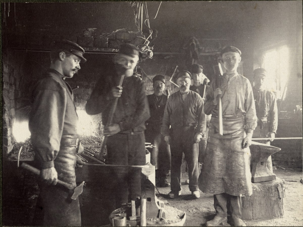 Schwarzes und weißes Foto einer Gruppe von Männern in einer Schmiede, jeder hält einen Hammer.