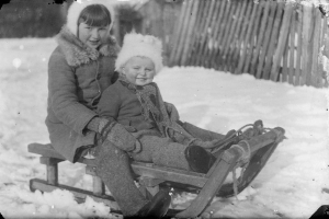 Ein altes Schwarz-Weiß-Foto von zwei Kindern in Mänteln und Mützen, die auf einem Schlitten in einer verschneiten Landschaft mit einem hölzernen Zaun im Hintergrund sitzen.