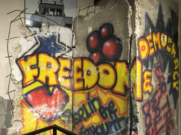 Eine Wand mit mutigem, schwarzem Graffiti, das "Freiheit und Demokratie" lautet, in einem Raum mit einem Geländer unten und einem Gebäude mit Fenstern im Hintergrund.