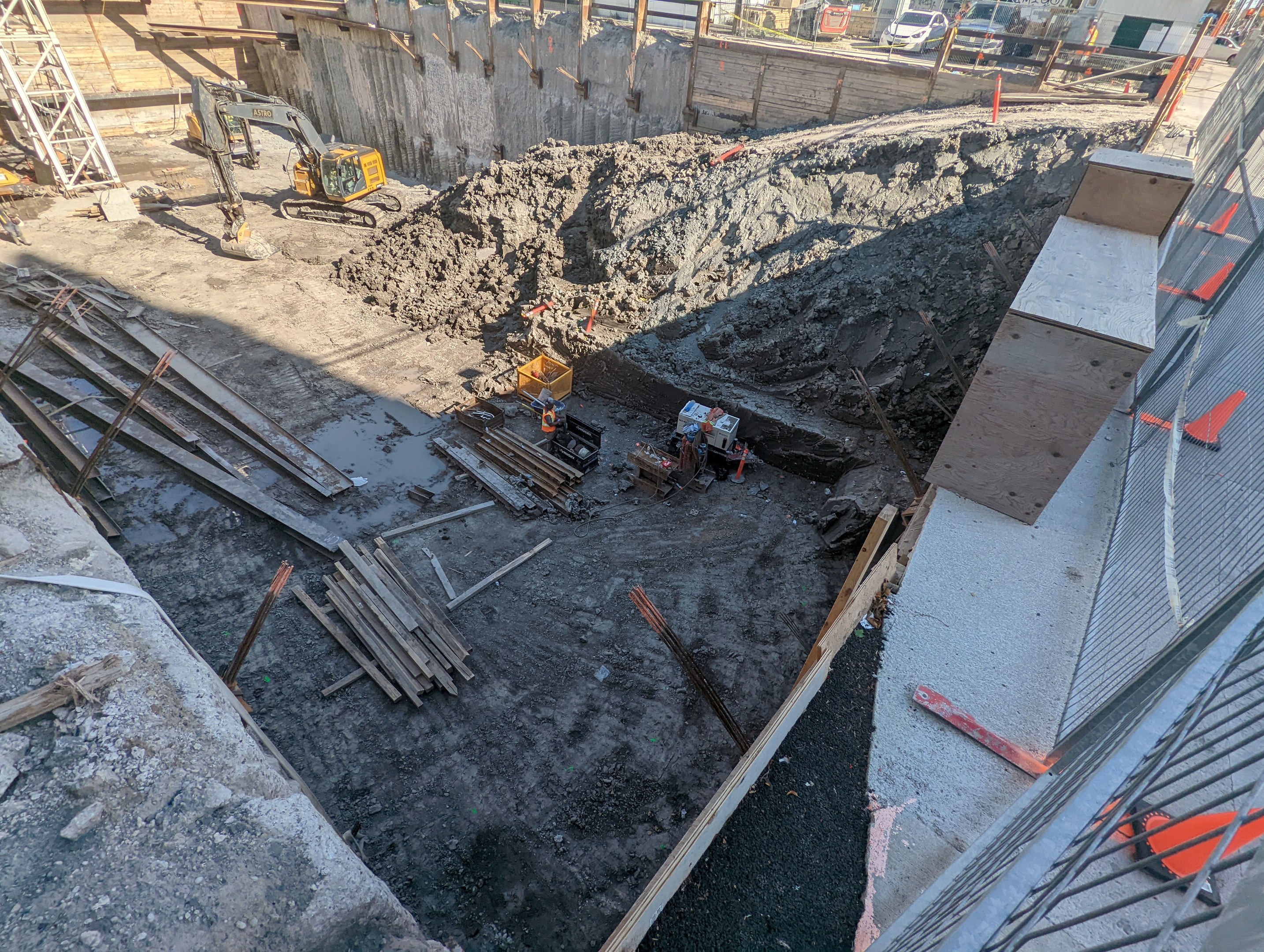 Baustelle mit einem großen Haufen Erde, umgeben von Kränen, Holzstangen und anderem Equipment, abgegrenzt durch einen Maschendrahtzaun, mit Gebäuden, Fahrzeugen, Pfählen, Bäumen und Himmel im Hintergrund.