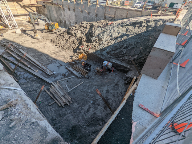Baustelle mit einem großen Haufen Erde, umgeben von Kränen, Holzstangen und anderem Equipment, abgegrenzt durch einen Maschendrahtzaun, mit Gebäuden, Fahrzeugen, Pfählen, Bäumen und Himmel im Hintergrund.