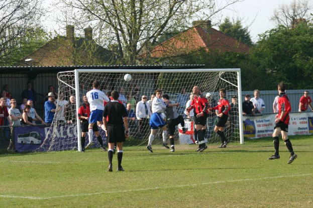 Fußballspieler sind in einem Spiel auf einem Feld mit einem Tornetz beteiligt, während Zuschauer dahinter stehen; Bäume und Häuser sind im Hintergrund sichtbar.