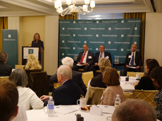 Eine Frau steht an einem Rednerpult mit einem Mikrofon und spricht zu einer Gruppe von Menschen, die an einem Tisch mit Wasserflaschen, Tassen und Papieren sitzen.