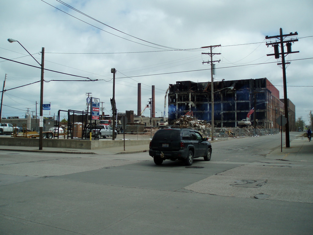 Ein Fahrzeug auf der Straße mit einer Laterne mit Kabeln auf der linken Seite und einer weiteren Laterne mit Kabeln auf dem rechten Gehweg, Gebäuden, anderen Fahrzeugen und Wolken am Himmel im Hintergrund.