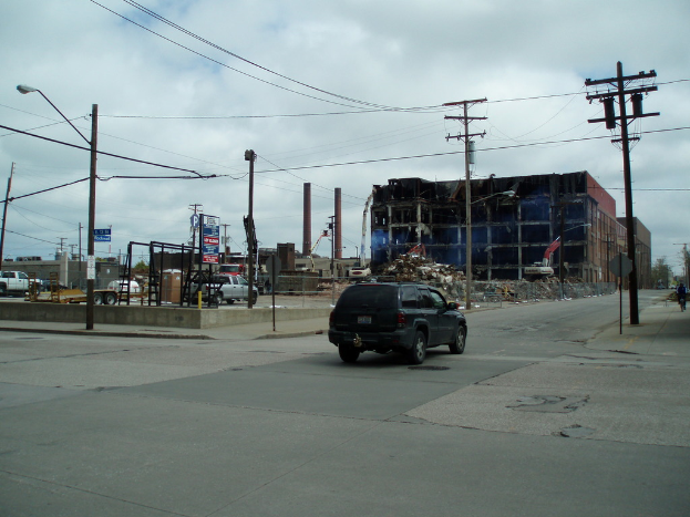 Ein Fahrzeug auf der Straße mit einer Laterne mit Kabeln auf der linken Seite und einer weiteren Laterne mit Kabeln auf dem rechten Gehweg, Gebäuden, anderen Fahrzeugen und Wolken am Himmel im Hintergrund.