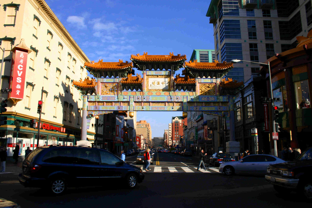 Eine Straße mit fahrenden Fahrzeugen, Fußgänger an einer Zebrastreifen, ein Bogen im Hintergrund, Gebäude auf beiden Seiten und eine Ampel unter einem klaren Himmel.
