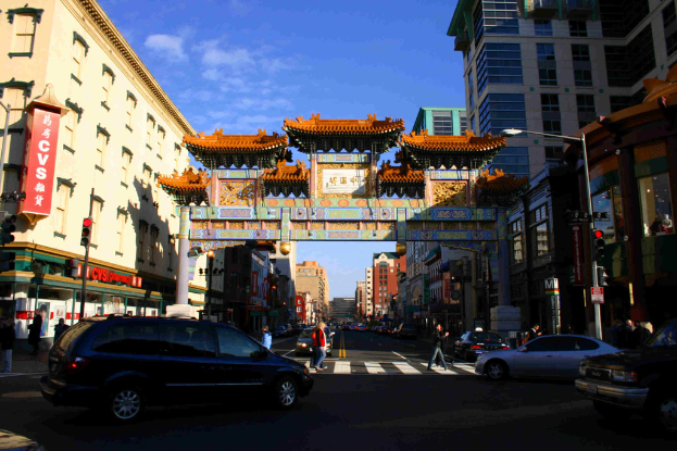 Eine Straße mit fahrenden Fahrzeugen, Fußgänger an einer Zebrastreifen, ein Bogen im Hintergrund, Gebäude auf beiden Seiten und eine Ampel unter einem klaren Himmel.