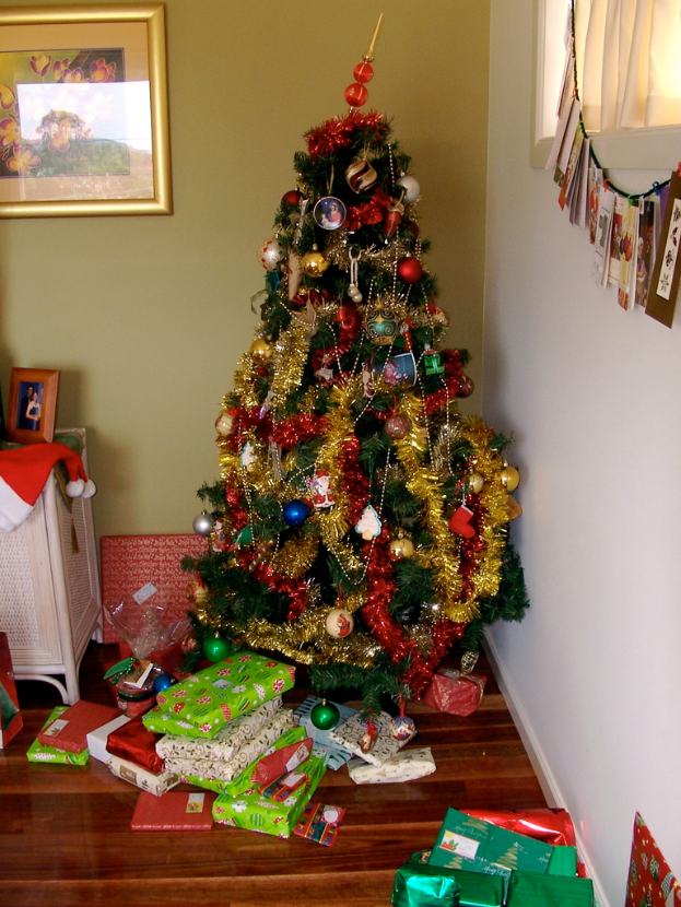 Ein geschmückter Weihnachtsbaum rechts mit Geschenken am Fuße, ein Foto Rahmen an der linken Wand und ein Vorhang am rechten Fenster.