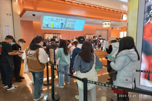 Eine Gruppe von Menschen steht an einem Restauranttresen Schlange, einige tragen Mützen und tragen Taschen, mit Stangen, die mit Bändern geschmückt sind, einem Bildschirm mit Text, Deckenlampen und Tischen mit verschiedenen Gegenständen im Hintergrund.