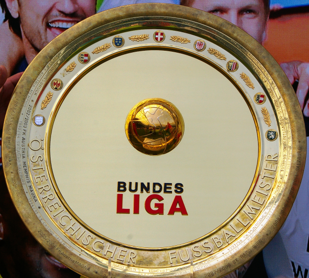 Ein Mann hält ein goldenes Schild mit der Aufschrift 'Bundesliga' hoch, mit einem Banner im Hintergrund mit drei Personen.