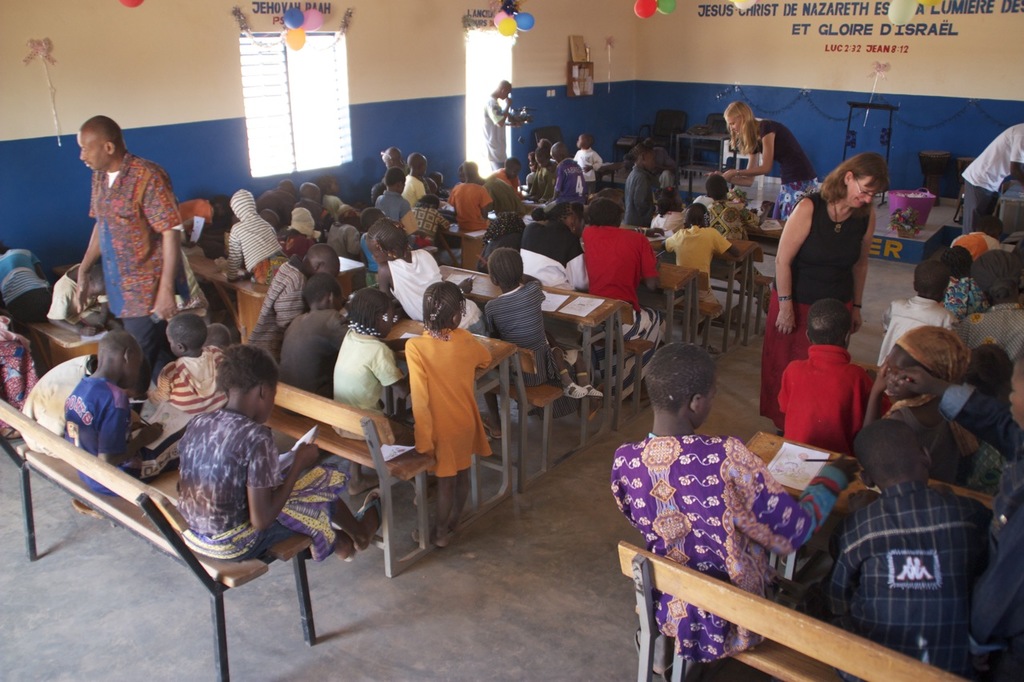 Kinder auf Bänken im Vordergrund mit Erwachsenen, die sie führen, Stühle, eine Tür, ein Fenster und ein Tisch im Hintergrund, Ballons und Text oben.