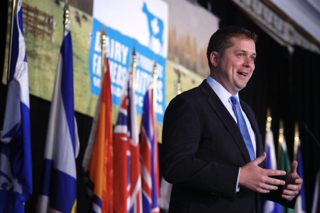 Ein Mann im Anzug spricht an einem Podium mit Mikrofonen, umgeben von Flaggen und einem Bildschirm, wahrscheinlich zu einer Regierungszahlung an die Milchindustrie.