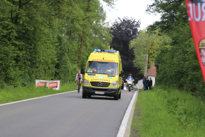 Ambulanz fährt auf einer Straße mit Fahrradfahrern nebenher, Gras und Bäume auf beiden Seiten, Häuser und Masten im Hintergrund unter einem klaren blauen Himmel.