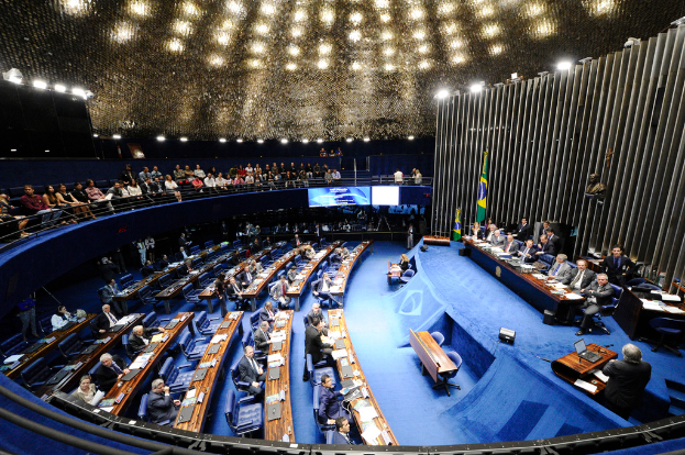 Eine Konferenzbühne mit sitzenden Personen, einem Tisch mit Papieren und Gegenständen, Fahnen und einer Skulptur im Hintergrund, mit Sitzplätzen vor der Bühne und hinter einem Geländer, beleuchtet von Deckenlampen.