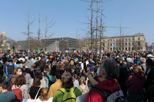 Eine große Menge Menschen steht vor einem Gebäude mit Fenstern, Bäumen und einem klaren Himmel, viele halten Schilder und tragen Täschchen, was auf eine Klimaprotestveranstaltung hindeutet.