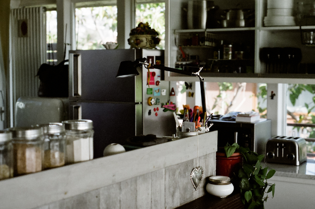 Ein unordentlicher Raum mit Flaschen, einem Topf, einer Pflanze, verschiedenen Gegenständen, einer Tischlampe, einem Kühlschrank mit Gegenständen darauf, Fenstern, einem Schrank voller Gegenstände und einer Plattform mit elektronischen Geräten.
