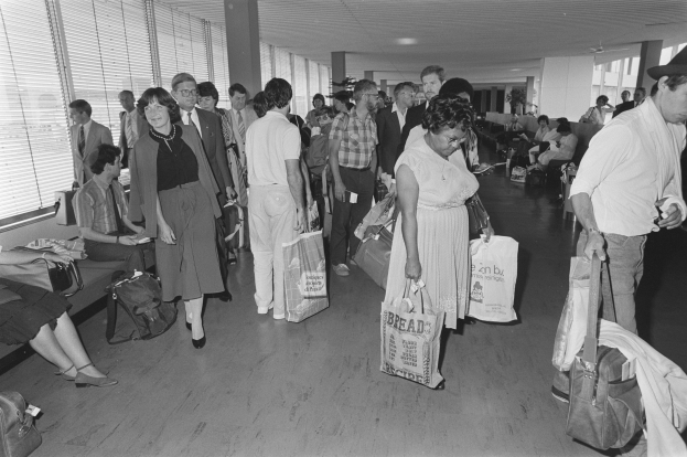 Schwarzes und weißes Bild von Reisenden in einem Flughafen, einige sitzen auf Bänken und andere stehen mit Gepäck, mit einem Mann und einer Frau, die Taschen in der Mitte nahe Fenstern mit Jalousien halten.