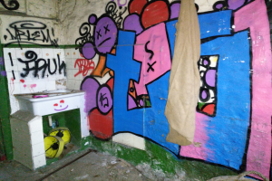 Toilette mit beschmierten Wänden, Waschbecken in der Ecke mit einem Tuch bedeckt und verstreute Gegenstände auf dem Boden.