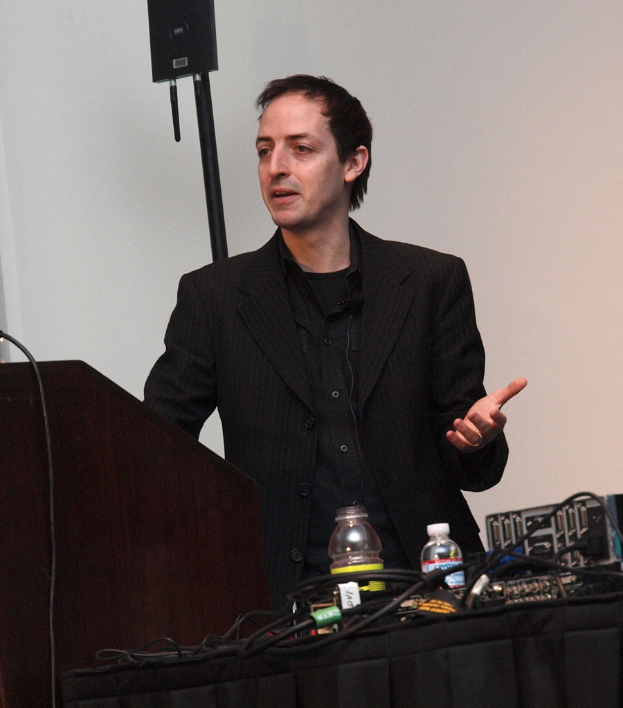 Ein Mann in einem Anzug steht neben einer Wand mit Flaschen und Kabeln.