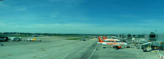Eine Flughafenszene mit Flugzeugen, Gebäuden, Polen, Fahrzeugen und Bäumen, einem Himmel oben, einer Straße unten und einem Zaun im Vordergrund.