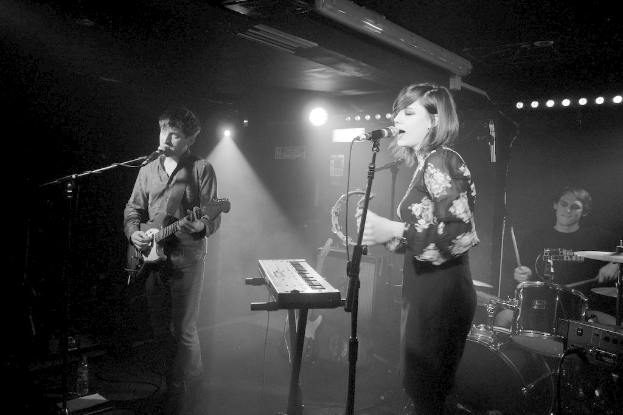 Ein Mann mit einer Gitarre und eine Frau, die beide in Mikrofone singen, mit einem Schlagzeuger im Hintergrund.