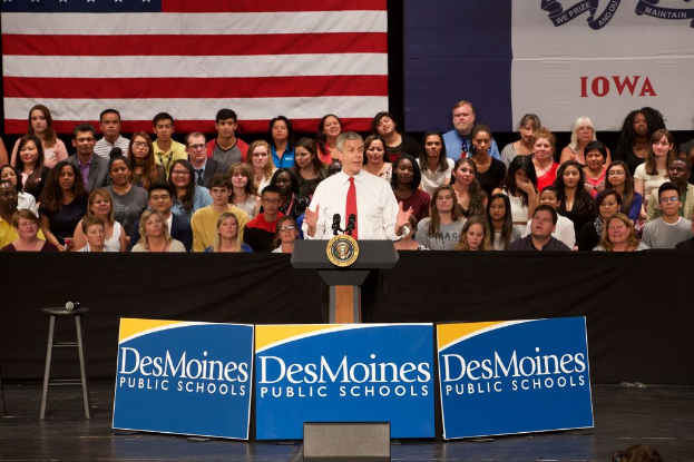 Ein Mann in einem weißen Hemd und roter Krawatte steht an einem Rednerpult mit drei Schildern, einem Hocker mit Mikrofon und einer sitzenden Menge im Hintergrund, mit einer Fahne und zusätzlichem Text.