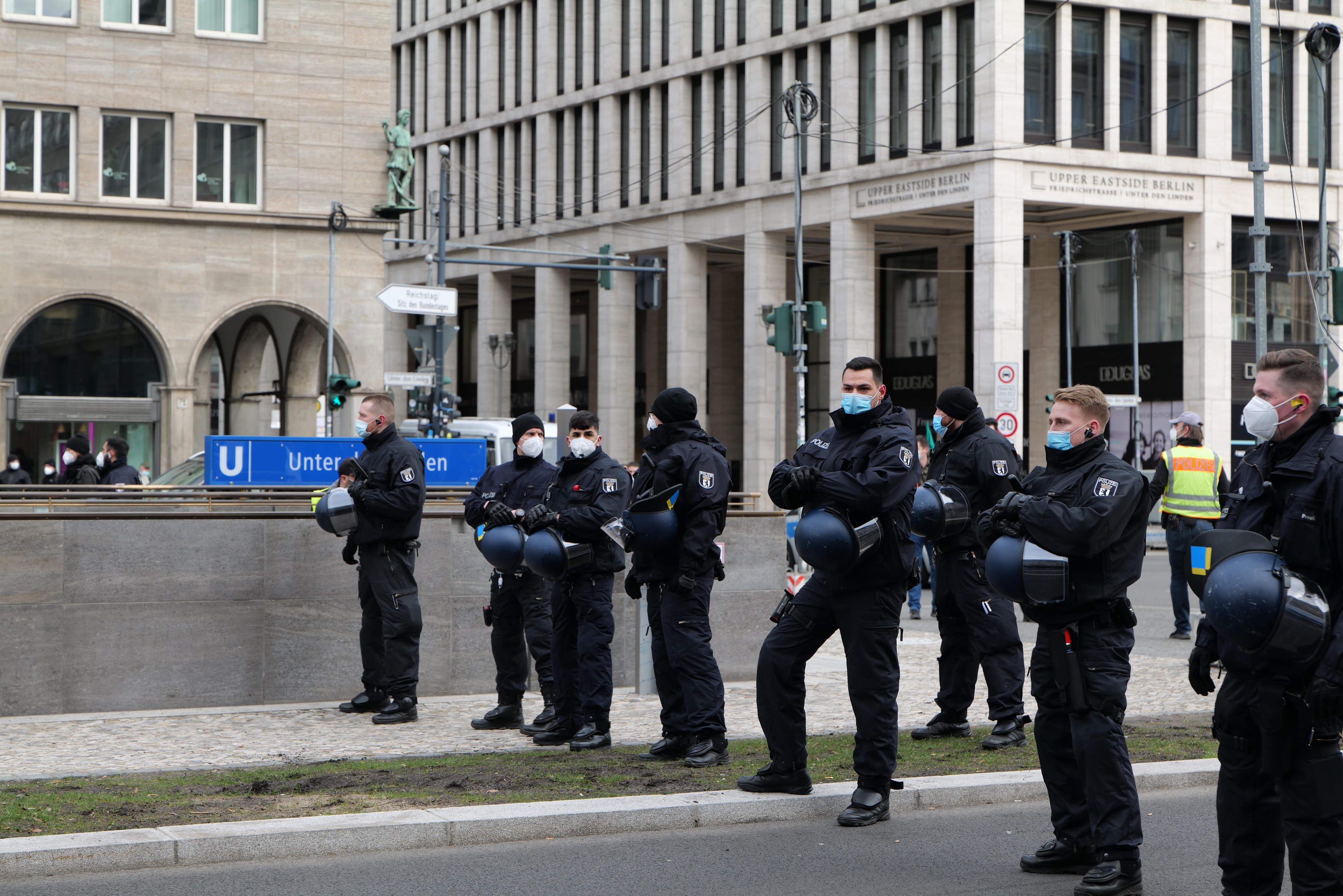 Polizeibeamte in schwarzen Uniformen und Masken stehen vor einem Berliner Gebäude mit Glasfenstern und Säulen, einige halten Helme, mit Laternen, Verkehrszeichen, Schildern mit Text, einer Statue und anderen Menschen im Hintergrund.