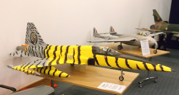 Verschiedene Flugzeugmodelle sind auf Tischen mit Etiketten angeordnet, ein Stuhl befindet sich auf der linken Seite und eine Wand im Hintergrund.