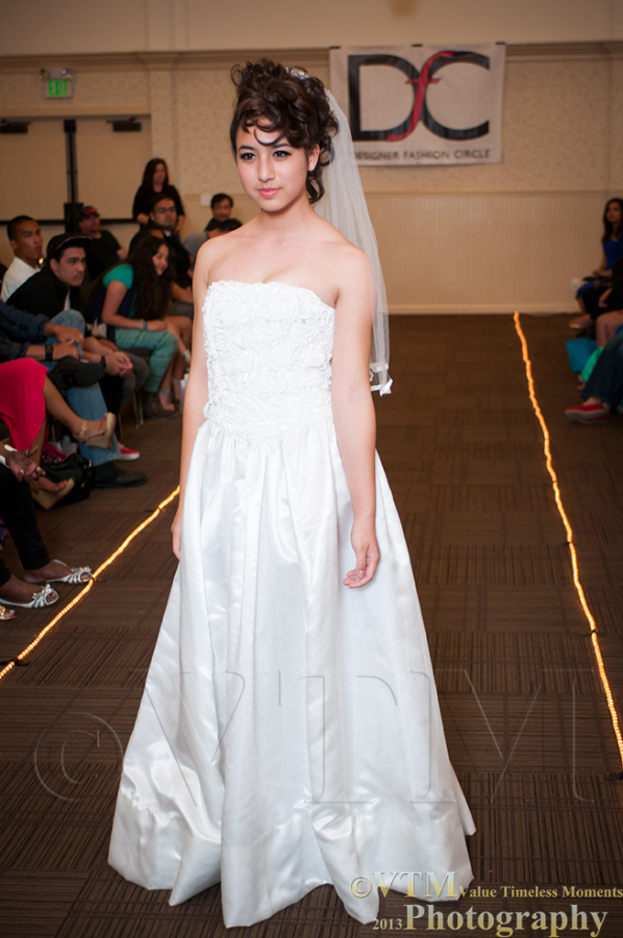 Eine Frau in einem weißen Kleid lächelt und posiert vor einem weißen Banner mit Text, umgeben von Menschen auf Stühlen und einem braunen Boden.