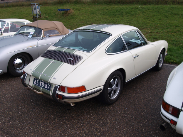 Weißer Porsche 911 auf einem grasbewachsenen Parkplatz mit anderen Autos, einem Zaun, Pflanzen, Bäumen, einem Gebäude und einer Tafel mit Text im Hintergrund geparkt.
