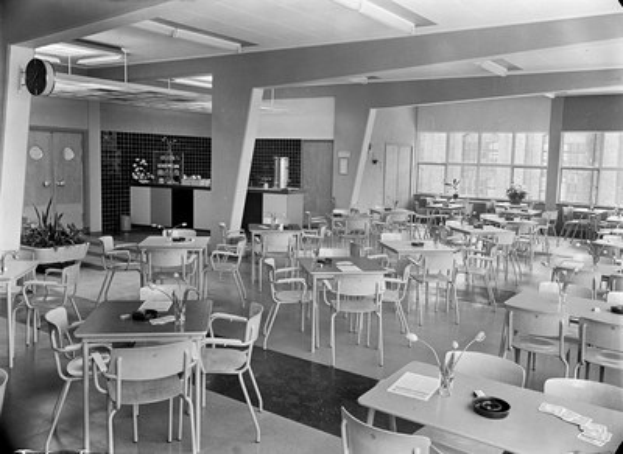 Schwarz-weißes Foto einer Cafeteria mit Tischen und Stühlen, Tellern, Besteck, Gläsern und Blumenvasen auf den Tischen und einer Wand mit Türen, Fenstern und Deckenlampen im Hintergrund.