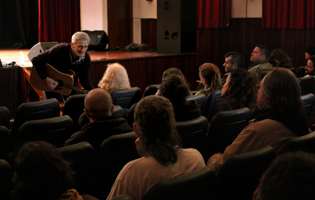 Ein älterer Mann in einem schwarzen Pullover spielt Gitarre auf einer Bühne mit Lautsprechern und einem Vorhang, während das Publikum auf Stühlen vor ihm sitzt.