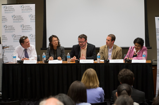 Ein Konferenzraum mit einem Tisch, der Namensschilder, Flaschen und ein Mikrofon enthält, fünf Personen, die darum herum sitzen, ein Podium und ein Banner links und ein Publikum davor.