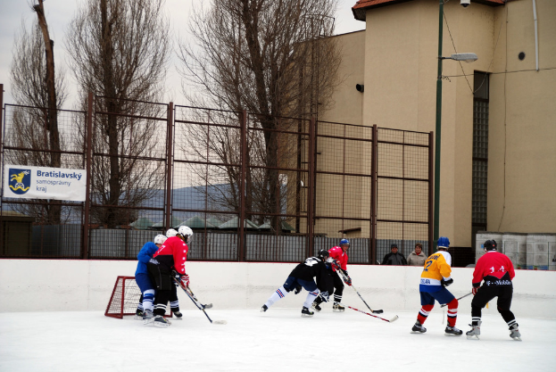 Menschen beim Eishockey-Spielen mit Himmel, Bäumen, Gebäuden, Straßenlaterne, Namensschild und Zäunen im Hintergrund.