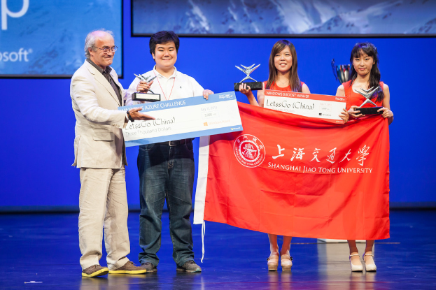 Vier Personen auf einer Bühne mit Auszeichnungen, Schecks und einer Fahne, die strahlend vor einem digitalen Bildschirm stehen.