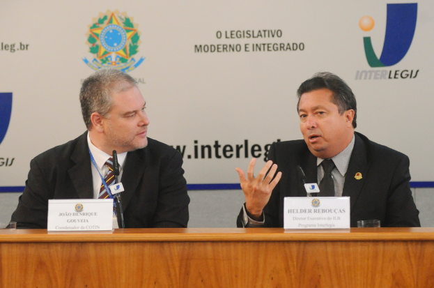 Zwei Männer in Anzügen sitzen an einem Tisch mit zwei Namensschildern, zwei Mikrofonen und Logos im Hintergrund.