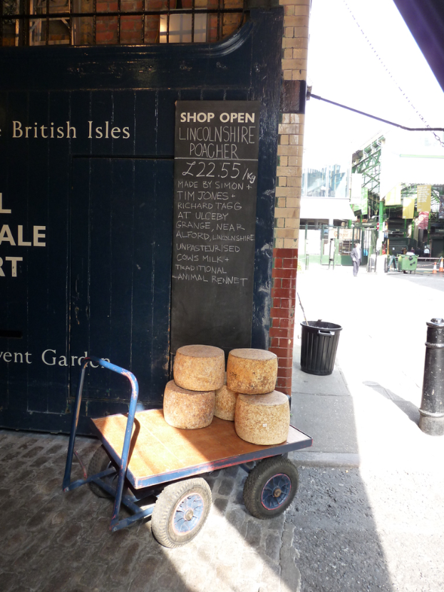 Ein Gepäckwagen mit Gegenständen darauf steht vor einer schwarzen Tür und einem schwarzen Schild mit Text, mit einem Gebäude, einem Müllcontainer und einem Pfahl in der Nähe, und Menschen mit Gebäuden und einer Treppe im Hintergrund.