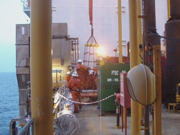 Ein Schiff mit Masten, Kisten und Geländern, das Menschen mit Helmen zeigt, mit Wasser im Hintergrund.