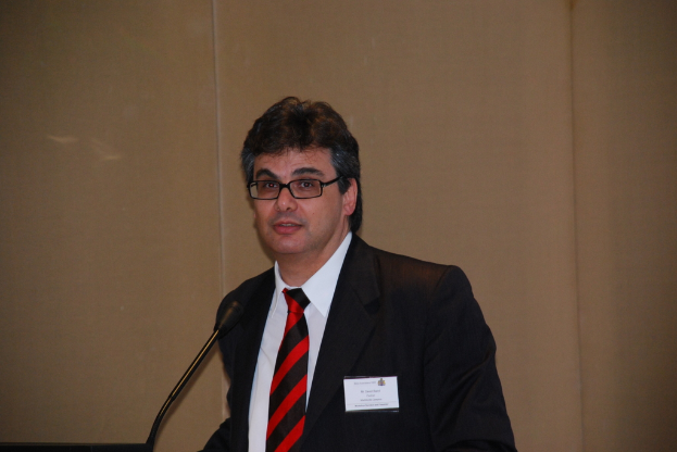 Eine Person in einem Anzug, die eine Marke und eine Brille trägt, lächelt und vor einem Mikrofon auf einem Ständer steht, mit einer Wand im Hintergrund.