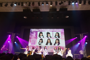 Eine Gruppe von Menschen, die auf einer hell erleuchteten Bühne performen, mit einer Menge darunter, mit einem Sänger mit Mikrofon und einem Musiker, der ein Instrument spielt, und einem großen Bildschirm im Hintergrund, der Bilder von BK48 anzeigt.