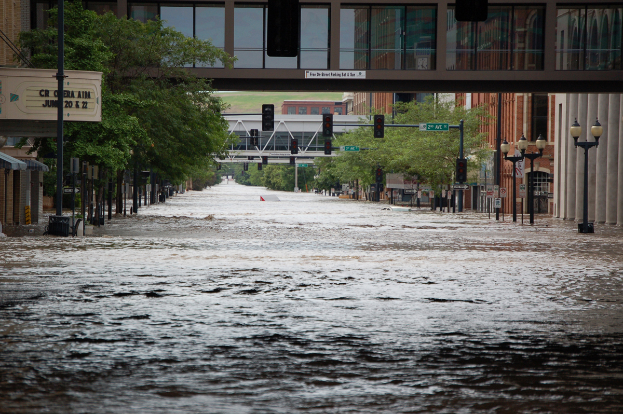 Eine überflutete Stadtstraße mit Wasser, das die Straße, Pfähle, Laternen, Schilder, Verkehrszeichen, Bäume, Gebäude und eine Brücke im Hintergrund bedeckt.