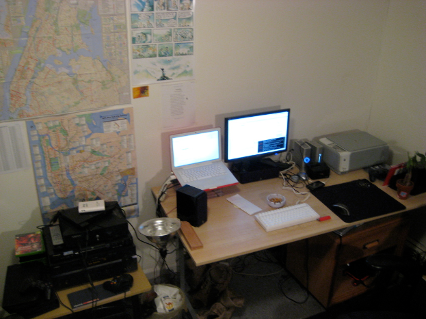 Ein Raum mit einem Tisch, auf dem ein Laptop, ein Lautsprecher, ein Monitor, eine Tastatur, ein Ball, eine Maus, ein Scanner und Papiere liegen, sowie ein weiterer Tisch mit Elektronikgeräten und Charts und Postern an der Wand dahinter.