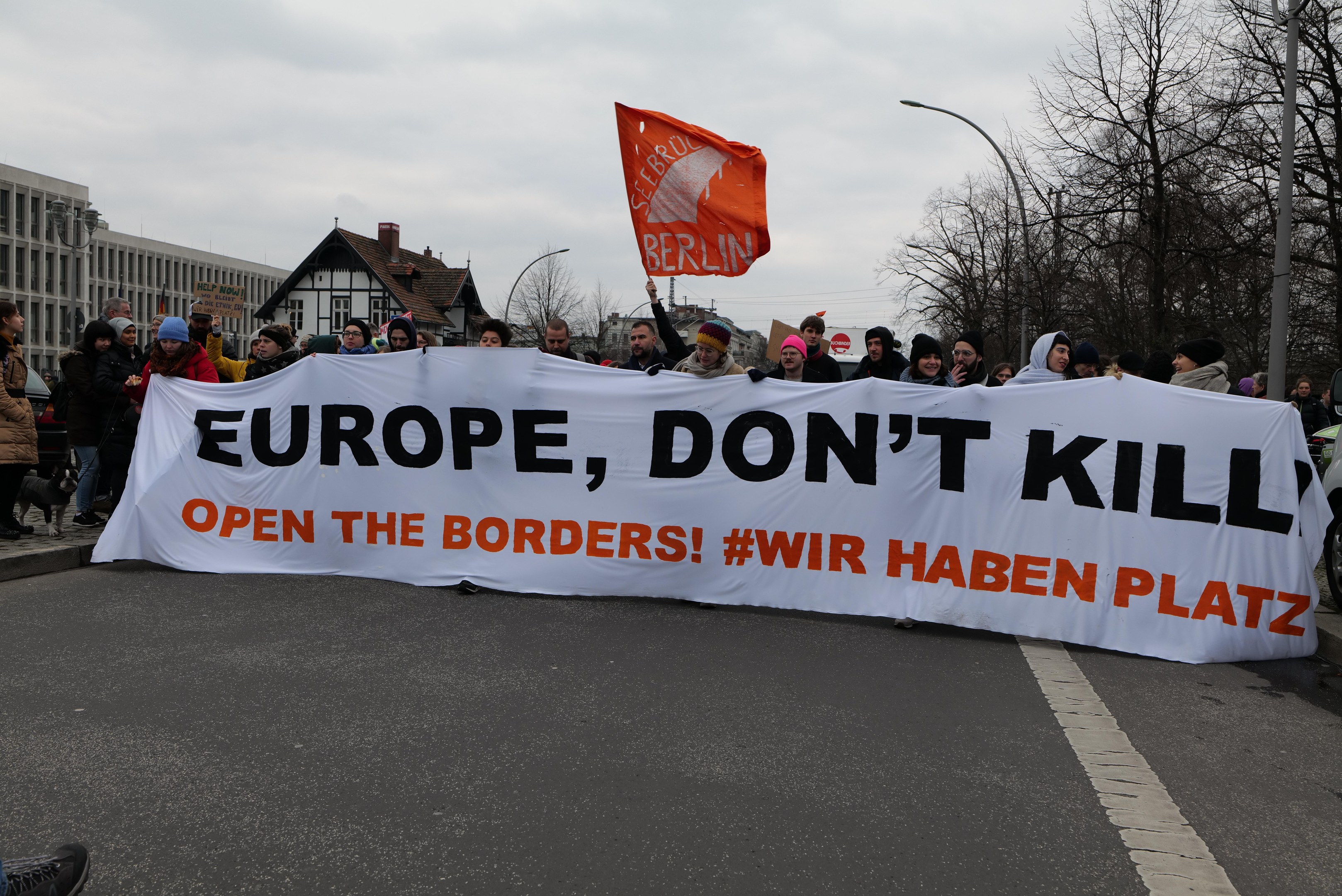 Gruppe von Menschen mit einem Banner "Europa, töte nicht Öffne die Grenzen" und einer Flagge auf einer Straße, mit einem Auto auf dem Gehweg und Gebäuden, Bäumen und Laternen im Hintergrund.