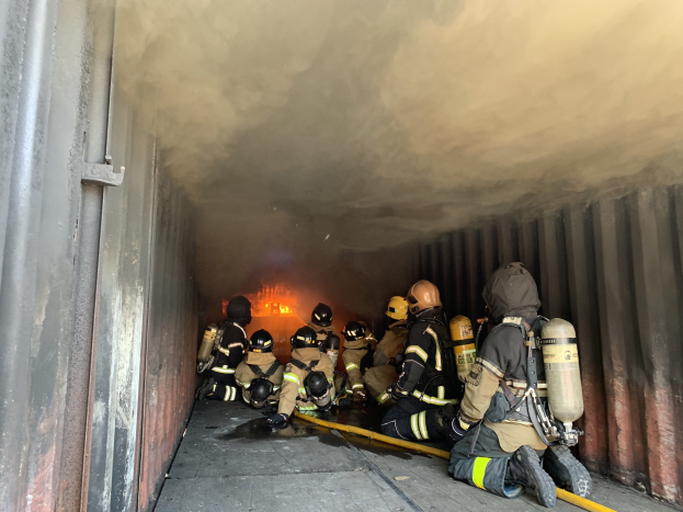 Feuerwehrleute in Schutzausrüstung arbeiten in einem raucherfüllten Tunnel, einer hält ein Rohr, mit sichtbarem Feuer und einer Wand auf der linken Seite.