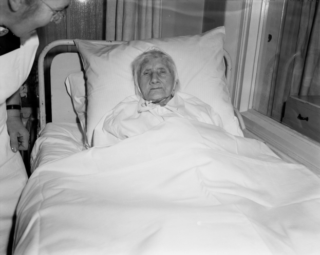 Ein älteres Weib liegt in einem Krankenhausbett mit einem weißen Laken und Kissen, während eine Krankenschwester neben ihr steht, mit einem Fenster mit Vorhängen im Hintergrund.