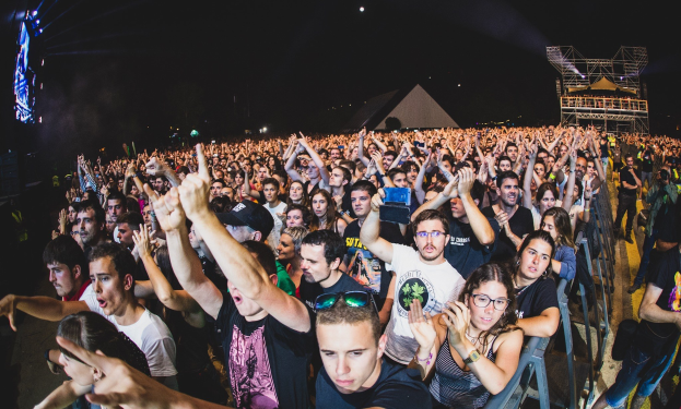 Menschenmenge auf einem Musikfestival mit erhobenen Händen, einige sitzen auf Stühlen und andere stehen, in der Nähe eines Metallzauns mit Gebäuden und Lichtern im Hintergrund.